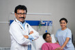 © WESTOCK - Rack focus shot, confident indian doctor standing with crossed arms in front of patient by looking camera at hospital - concept of professional occupation, expertise and successful treatment