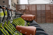  Close-up Of A Row Of Green Bicycles With Brown Leather Seats And Silver Studs, Parked In A Bike Rack. The Focus Is On The First Bicycle's Seat. The Backdrop Features A Wooden Fence And A Building.