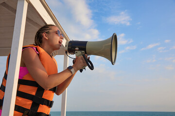 Young female lifeguard in an orange life jacket shouts an announcement into the loudspeaker for beachgoers. Woman in sunglasses broadcasts into the horn from the rescue tower