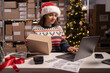 Employee in retail shop's warehouse in Santa hat. Small Business Owner working on laptop, preparing and packing parcels for delivery in Christmas Eve.