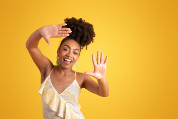 Wall Mural - Smiling millennial black lady in dress making frame, lens sign with hands