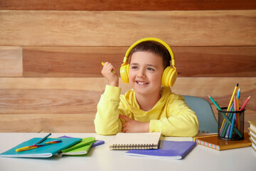 Poster - Cute little boy in headphones doing homework at table near wooden wall