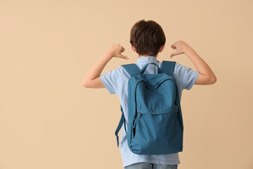 Little boy pointing at backpack on beige background, back view