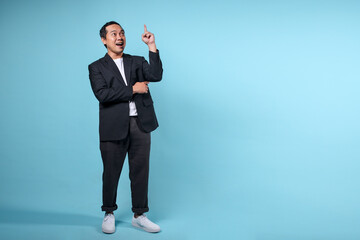 Full length portrait of happy smiling Asian businessman pointing finger up isolated over blue background