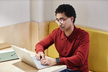 Wall Mural - Portrait of Middle Eastern young man wearing glasses and using computer at desk in booth with electric lighting, copy space