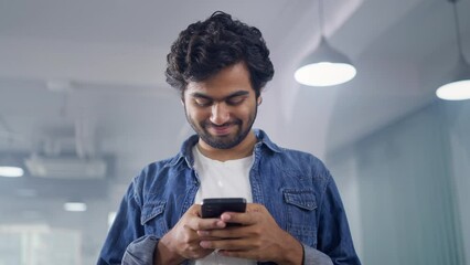 A young Indian Asian modern male or hipster man is standing indoors using a mobile phone to type a text message or post a comment on a social media platform looking into the camera and smiling.
