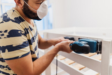 man rubs wooden child bed with power sander or grinder removing old paint