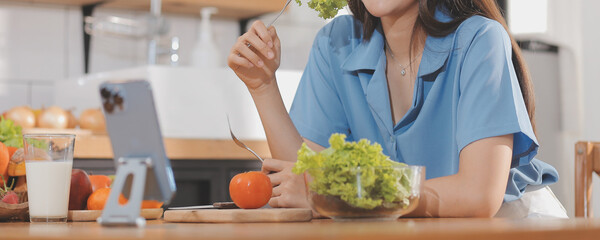 A young woman with a beautiful face in a blue shirt with long hair eating fruit sitting inside the kitchen at home with a laptop and notebook for relaxation, Concept Vacation.