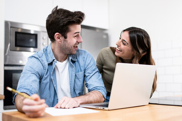 young couple planning budget together at home writting notes to paper and looking a laptop. two cheerful young adults sitting on the kitchen. finance concept