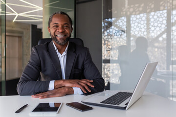 Portrait of successful mature adult businessman, man with crossed arms smiling and looking at camera, african american boss financier sitting at table with laptop.