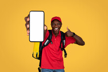 Glad Young African American Delivery Man In Uniform With Big Backpack Show Thumb Up Sign, Phone With Blank Screen