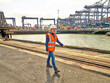 © Connect Images - Female dock worker walking by dock with cargo cranes in background, Felixstowe, England.
