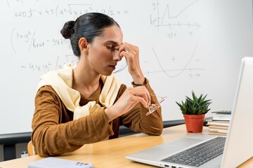 Tired business woman working inside modern office or classroom, worker rubbing eyes with glasses off, Latin American woman with black hair using laptop for work