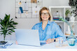 © okrasiuk - Smiling mature businesswoman with laptop at her office workplace, confident middle-aged experienced senior female professional, CEO of company looking at camera. Creative entrepreneur manage business.