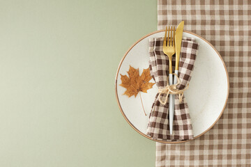 Autumn-themed table decor inspiration. Top view arrangement of plate, cutlery, napkin, tablecloth, autumn maple leaf on pastel olive background with empty space for advert or message