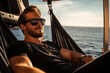 A man in a black tshirt and sunglasses chilling in a luxurious hammock strung up in the cabin of the superyacht looking out at the