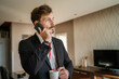 © Miljan Živković - one man adult caucasian businessman wear suit in hotel room while taking a brake or prepare for work hold cup of coffee and smartphone making a phone call talk real people copy space