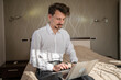 © Miljan Živković - One man adult caucasian businessman sit on the bed in hotel room work on his laptop computer prepare for meeting or remote online conference real person copy space wear white shirt