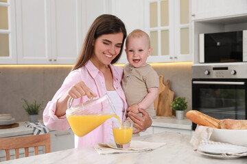 Canvas Print - Happy young woman holding her cute little baby while pouring juice into glass in kitchen