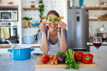 Wall Mural - Photo of young woman smiling with green pepper slices while cooking salad with fresh vegetables in kitchen interior at home