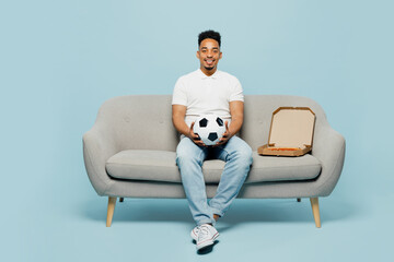 Wall Mural - Full body smiling happy cheerful young man fan in t-shirt cheer up support football sport team hold soccer ball sit on grey sofa looking camera watch tv live stream isolated on plain blue background