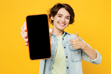 Young happy woman wear green t-shirt denim shirt casual clothes hold in hand use close up mobile cell phone with blank screen workspace area show thumb up isolated on plain yellow background studio.