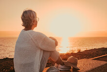 Back view of elderly mature woman sitting alone face the sea at sunset light looking at the horizon. Senior gray-haired woman enjoying freedom and beautiful seascape in vacation