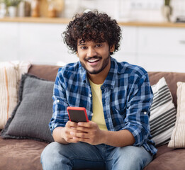 Happy young Asian man with curly hair using mobile phone at home, messaging or browsing social networks while relaxing sitting on sofa looking at camera and smiling friendly