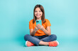 © deagreez - Photo of cheerful pretty girl pupil girl dressed stylish clothes sitting floor playing game iphone isolated on cyan color background