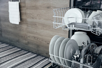 Closeup of opened automatic stainless built-in dishwasher machine inside modern home kitchen with clean utensil. White plates, dishes, cups, metal forks and knifes. Household, housekeeping, housework