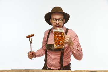 Wall Mural - Bavarian mustache man wearing traditional fest outfit holding hot fried sausage and looking at huge glass of beer. Oktoberfest concept