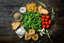 Italian Food Ingredients, Fresh Mozzarella, Bruschetta, Raw Tagliatelle And Fusilli, Basil Leaves, Tomatoes, Garlic, Arugula, Olive Oil And Pepper On A Dark Wooden Background