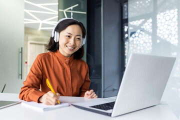 Asian business woman with headphones watching online training course at workplace, woman writing information happy and satisfied with the results of professional development