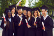 © Studio Romantic - Group of several university students on graduation day. Happy diverse young friends in graduate hats and gowns standing in green campus yard, holding diploma scrolls, looking at camera and smiling