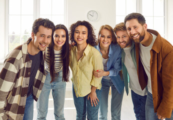 Group of happy smiling funny friends students or colleagues in casual clothes standing together and looking cheerful and positively at the camera. Portrait of friendly guys and girls indoors.