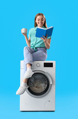 Canvas Print - Pretty young woman sitting on washing machine, reading book and drinking tea against blue background