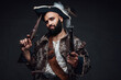 © Fxquadro - Attractive pirate with a black beard, wearing a vest and hat, holding two muskets against a dark textured wall