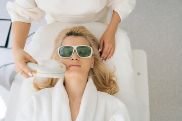 Closeup high-angle view of young woman client in protective glasses getting photo rejuvenation procedure at cosmetology clinic. Concept of technology hardware for anti age-related changes of face.