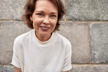 Wall Mural - Close-up portrait of attractive mature woman against the background of a stone wall. Middle-aged brunette in a white t-shirt smiles happily. Active lifestyle for the elderly.