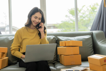 A young Asian small business entrepreneur starts using a smartphone and laptop computer to check orders from the web, owns a work at home online seller prepares product parcel packs for delivery.