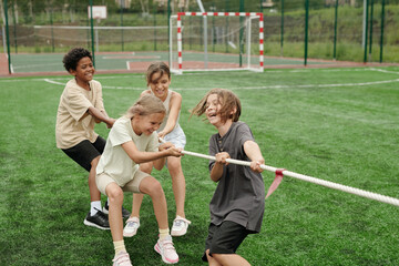 Cheerful schoolkids in activewear laughing while pulling rope during sports competition with another team on green football field at stadium