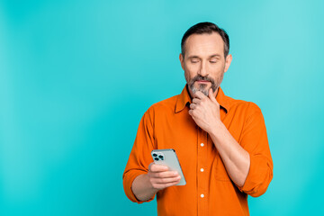 Canvas Print - Portrait of ponder pensive man dressed orange shirt look at smartphone read post arm on chin isolated on turquoise color background