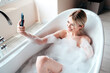 © halayalex - Young beautiful sexy woman having fun while lying in bathtub full of foam at home. Charming smiling model relaxing in luxury bath interior. Female holding smartphone, taking selfie photos, video call