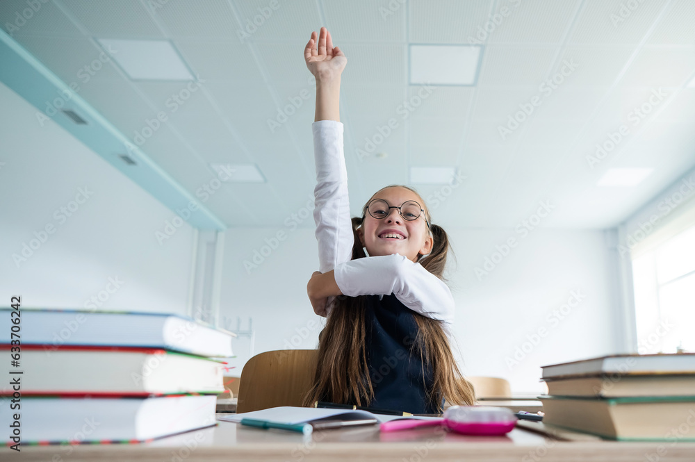 Caucasian girl with two ponytails sits at her desk and raises her hand ...