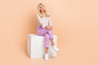 © deagreez - Full length photo of lovely pretty lady wear stylish outfit sitting white cube watching empty space isolated on beige color background