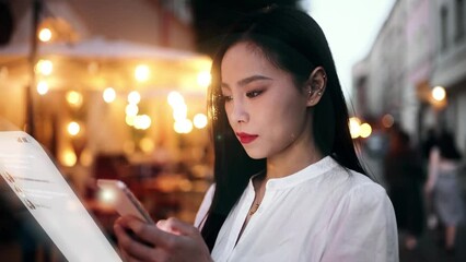 Wall Mural - A beautiful young Asian woman is typing a message on an augmented reality device while standing on an evening city street. The concept of social networks, futurism and communication