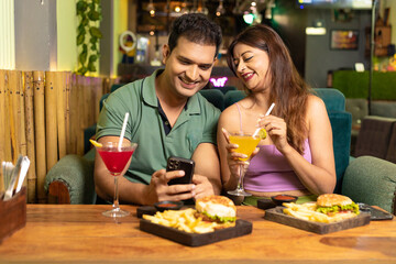 Wall Mural - Indian Couple Looking in smartphone while enjoying food and drink at restaurant.