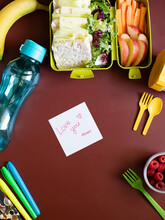 School Lunch Box With Sandwiches, Carrot Sticks, Apple, Banana, Lettuce, Hummus And Raspberries. Healthy School Lunch Concept. Burgundy Background And Note With Text - Love You Mom. Top View 