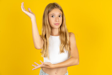 blonde kid girl wearing white T-shirt over yellow studio background pointing aside with both hands showing something strange and saying: I don't know what is this. Advertisement concept.