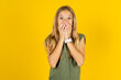 Vivacious blonde kid girl wearing green T-shirt over yellow studio background , giggles joyfully, covers mouth, has natural laughter, hears positive story or funny anecdote